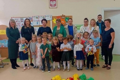 Dzieci stoją w dwóch rzędach, w pierwszym Dzieci pasowane, na głowach mają kolorowe czapki, w ręku trzymają dyplomy i upominki. Z prawj strony stoi wychowawczyni. W tle widać kolorowe tablice z dekoracją.