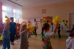 Dzieci bawią się w kole do piosenki, w rękach trzymają kolorowe pompony z bibuły. W tle widać kolorowe tablice.