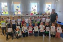 Grupa dzieci i kobieta stoją na tle okien trzymając w dłoniach kartki.