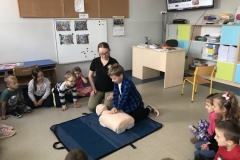 Chłopiec w niebieskiej koszuli w kratkę klęczy przed manekinem uciskaĵąc jego klatkę, podczas gdy dookoła niego siedzą dzieci.