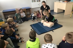 Grupa dzieci siedzi na podłodze w kółku, a dziewczynka klęczy w środku i uciska klatkę piersiową manekina.