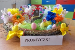W płaskim koszu z wikliny ułożone są różne elementy dekoracyjne o charakterze wielkanocnym. Na środku jest brązowa, filcowa kurka, z czerwonym dziobem             i grzebykiem oraz zielonymi skrzydełkami. Za nią ułożone są tekturowe pisanki ozdobione kwiatkami i motylkami. Na brzegach rozłożone cztery białe króliczki          z materiału. Po bokach bibułowe kwiaty. W środku przed kurą widać kolorowe małe jajeczka. Koszyk ozdobiony dwiema żółtymi kokardami do których przyczepione są małe żółte kurczątka. Z przodu napis czarnymi drukowanymi literami: Promyczki.
