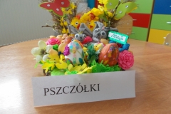 Na podkładce ozdobionej zieloną bibułą znajdują się elementy dekoracyjne o charakterze wielkanocnym. W środkowej części widoczne są dwa szare króliczki, przed nimi ozdobione styropianowe pisanki. Za nimi widać dwa żółte kurczaczki. Po bokach na gałązkach palmowych przypięte są dwa tekturowe motyle: czerwony i zielony. Całość ozdobiona kwiatkami z bibuły.Po prawej stronie mały niebieski  napis na białym tle: Alleluja. Z przodu napis czarnymi drukowanymi literami: Pszczółki.