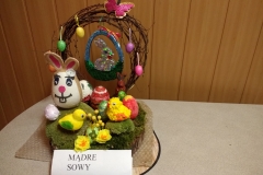 Na drewnianej podkładce ozdobionej zielonym mchem znajduje się beżowo - biały królik wykonany na styropianowym jajku, obok ozdobione pisanki ze styropianu,  żółta kaczuszka i kurczak oraz drobne kwiaty z bibuły. W tyle zwinięta w koło wiklina, na niej zawieszone malutkie kolorowe jajka, motyl oraz wycięty z kartonu kształt koszyczka z królikiem, ozdobiony srebrnym, błyszczącym materiałem.