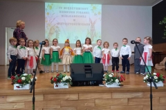 Grupa dzieci  stoi na scenie tworząc półkole. W tle biała ściana.