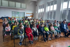 Dzieci siedzą na krzesłach w kilku rzędach.  Ubrane w kolorowe stroje.  W tle widać  duże okna.