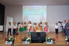 Dzieci w kolorowych strojach stoją na scenie. W tle jasna ściana.