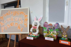 Na szarej tablicy widnieje napis Międzygminny Konkurs Pisanki. Obok stoi biały wielkanocny zajaczek oraz kolorowe jajka.