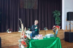 Na środku stoi stolik nakryty zielonym materiałem. Za nim  pani ubrana w ciemny strój. W tle brązowa kurtyna.