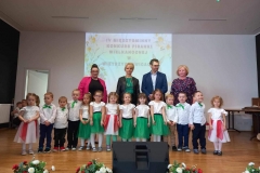 Dzieci w kolorowych strojach stoją na scenie tworząc półkole.  Za nimi trzy pani i jeden pan.