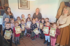 Grupa dzieci stoi  w dwuszeregu na tle jasnej ściany. Każdy trzyma w rękach dyplom w kształcie prostokąta.