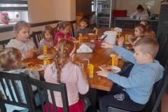 Wokół długiego drewnianego stołu, na ciemnych krzesłach siedzi 15 dzieci. Przed nimi stoi pani w dżinsowej koszuli, z boku 2 panie. Na stole leżą okrągłe placki, stoją 2 wiadereczka z żółtym serem, 2 pojemniki z pieczarkami, sos pomidorowy i kukurydza.