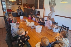 Dzieci siedzą przy prostokątnym brązowym stole. Obok stołu stoi pani w jasnym sweterku.