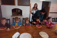 Pięcioro dzieci siedzi przy stoliku, dwie panie  stoją  za nimi ściana na której  wiszą dwie tablice.