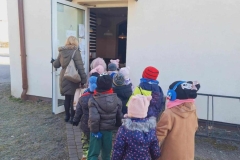 Dzieci w parach idą po chodniku z szarej kostki. Ubrane są w ciepłe kurtki, czapki, rękawiczki i szaliki. Pani w ciemnozielonej kurtce z szarą torebką na ramieniu otwiera drzwi do budynku. Nad drzwiami napis " Pizzeria".