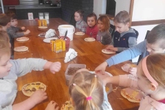 Duża grupa dzieci siedzi przy stoliku.  Przed nimi leżą okrągłe placki.