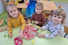 Dzieci podzielone na grupy siedzą przy 3 stolikach. Przed nimi pizza z szarego papieru. Dzieci naklejają na nią składniki: białe grzyby, czerwone pomidory i salami, fioletową cebulę, żółty ser.