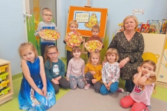 Na podłodze siedzi 6 dziewczynek. Za nimi stoi 3 chłopców. W rękach trzymają papierowe pizze, na których dominuje kolor czerwony i żółty. Obok nich na krzesełku siedzi pani. W tle tablica z napisem Dzień Pizzy i obrazem kawałka pizzy.