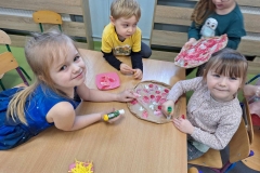 Dzieci podzielone na grupy siedzą przy 3 stolikach. Przed nimi pizza z szarego papieru. Dzieci naklejają na nią składniki: białe grzyby, czerwone pomidory i salami, fioletową cebulę, żółty ser.