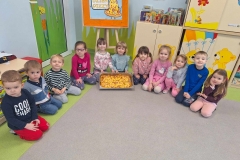 Grupa dzieci siedzi na szaro- zielonej podłodze. 2 dziewczynki trzymają w rękach blaszkę z upieczoną pizzą. Za nimi korkowa tablica z napisem Dzień Pizzy oraz szafka, na której stoją książki.