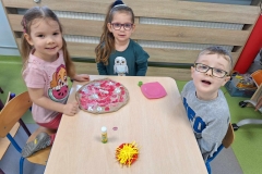 Dzieci podzielone na grupy siedzą przy 3 stolikach. Przed nimi pizza z szarego papieru. Dzieci naklejają na nią składniki: białe grzyby, czerwone pomidory i salami, fioletową cebulę, żółty ser.