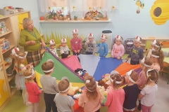 Dzieci wraz z panią stoją tworząc koło. Przedszkolaki trzymają kolorową chustę  animacyjną. W tle widać  dwa okna i szafki.