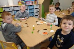 4 chłopców siedzi przy stoliku. Przed nimi leżą papierowe koła w różnych kolorach oraz klej. Chłopcy kleją głowę misia. W tle szufladki na przybory szkolne.