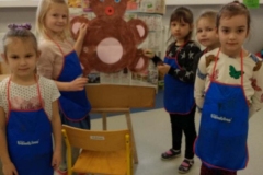 Na prostokątnej sztaludze wisi namalowany obrazek misia. Jest on w kolorze brązowym. Ma niebieskie oczy, czerwoną buzię różowy brzuszek. Obok sztalugi stoi 5 dziewczynek w niebieskich fartuszkach i krzesło.