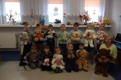 7 dzieci stoi w sali przedszkolnej. Przed nimi na podłodze siedzi 5 dzieci i pani w seledynowym swetrze. Wszyscy trzymają przed sobą misie w różnych kolorach. W tle okno z białymi firankami. Z boku na stoliku stoi komputer.