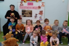 Sala zabaw. Grupa dzieci ubrana na kolorowo . 7 dzieci siedzi na krzesełkach , 3 dzieci siedzi na podłodze . Każde dziecko trzyma na kolanach swojego misia . Z prawej strony trzy misie ; jeden bardzo duży , a dwa mniejsze ułożone na podłodze- duży miś oparty o krzesełko. Za dziećmi postać dorosła- wychowawczyni ubrana na czarno trzyma małą pluszową zabawkę. W tle duża biała tablica z rysunkami misiów i napisem Dzień Pluszowego Misia na prostokątnych różowych paskach papieru .Powyżej czerwona tablica z sylwetami żółtych pszczółek.