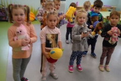 Grupa dzieci stoi w luźnej rozsypce przytula swoje misie. Za dziećmi widać część stolika i szafki z kącika kuchennego . W tle błękitna ściana z kolorowym nadrukiem drzewa i pszczółki oraz fragmenty dwóch okien.