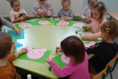 Grupa dzieci siedzi na krzesełkach przy okrągłym zielonym stoliku. Dzieci trzymają w ręce kleje i naklejają na prostokątne zielone kartki wycięte z papieru różowe koła ; jedno duże i dwa mniejsze układając głowę misia.