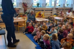 Dzieci siedzą na podłodze na niebieskich cienkich materacach. Przed nimi stoi policjant w granatowej koszuli i spodniach. W tle widać  okna.