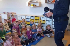 Grupa dzieci siedzi na podłodze. Przed nimi stoi policjalnt w granatowym stroju. W rękach trzyma policyjną czapkę.