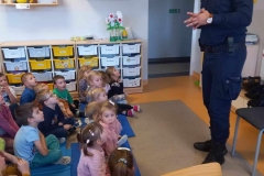 Grupa dzieci siedzi na podłodze. Przed nimi stoi policjant w służbowym granatowym stroju. W tle widać szafkę z białymi i żółtymi szufladami.