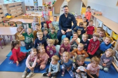 Dzieci siedzą na podłodze. Wśród nich na krześle siedzi policjant. W tle widać szafki.