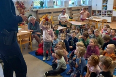 Grupa dzieci siedzi na podłodze. Przed nimi stoi policjant. W tle widać okna.