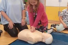 Dziewczynka w sukience i różowym sweterku klęczy nad manekinem i układa ręce na jego klatce piersiowej.