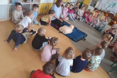 Grupa dzieci siedzi dookoła kobiety w niebieskiej bluzce i manekina.