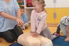 Dziewczynka w różowym sweterku klęczy nad manekinem obok kobiety w niebieskiej bluzce.