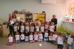 Grupa dzieci i dwie panie stoją na tle szafy i trzymają w dłoniach kartki.