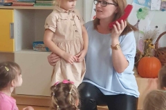 Kobieta w niebieskiej bluzce trzyma czerwony telefon i tłumaczy coś stojącej obok dziewczynce w kremowej sukience.