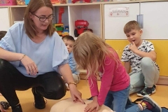 Dziewczynka w długich blond włosach i różowej bluzce klęczy nad manekinem i uciska jego klatkę piersiową.