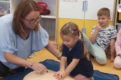 Mała dziewczynka z warkoczykami uciska klatkę piersiową manekina leżącego na ziemi.