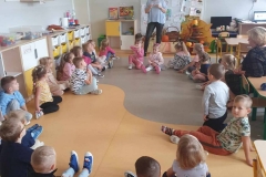 Grupa dzieci siedzi w kółku na podłodze i słucha kobiety w niebieskiej bluzce.