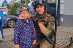 dziewczynka w kolorowej kurtce stoi obok kucającego obok niej żołnierza.