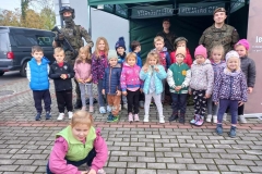 Dzieci ubrane w kolorowe stroje, stoją w dwóch szeregach. Za nimi dwóch żołnierzy w zielonych mundurach.