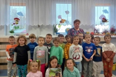 Trzy dziewczynki siedzą na podłodze. jedna z nich  trzyma dyplom. Pozostałe dzieci stoją w dwóch rzędach. W tle widać okno.