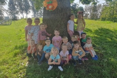 Jedenaścioro dzieci kuca na ziemi, pięcioro stoi. Przedszkolaki skupiły się wokół durzego pnia.