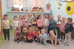 Sześcioro dzieci kuca na podłodze, za nimi dwanaścioro stoi wraz z panią. W tle widać dwa okna.